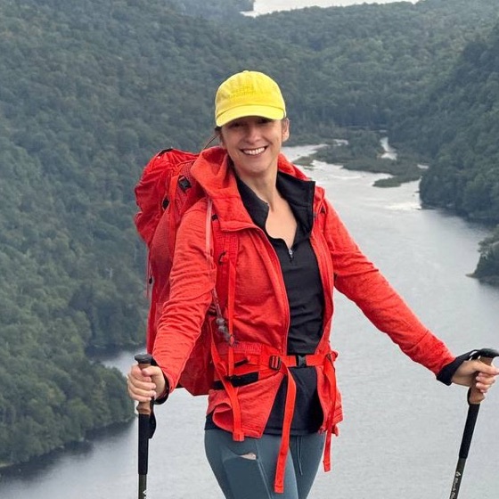Olga Manakina in a red jacket and yellow cap on a mountain hike, a lake behind her